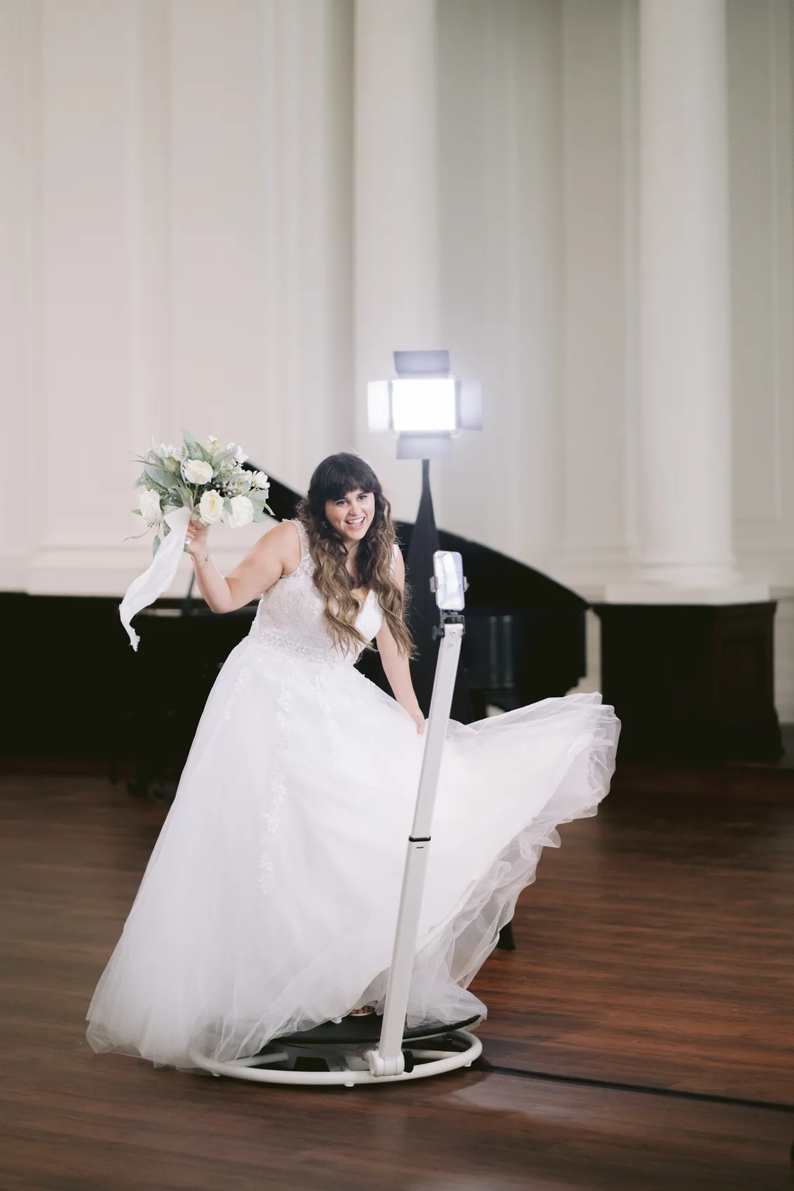 A bride sweeping her tulle skirt around her as she steps off the spinning platform, a grand piano behind her in a bright ballroom