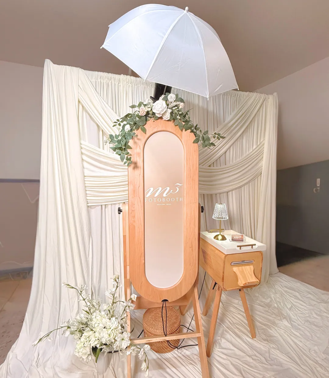 A wood framed mirror booth standing under a white umbrella, dressed with white roses and eucalyptus beside cream draping