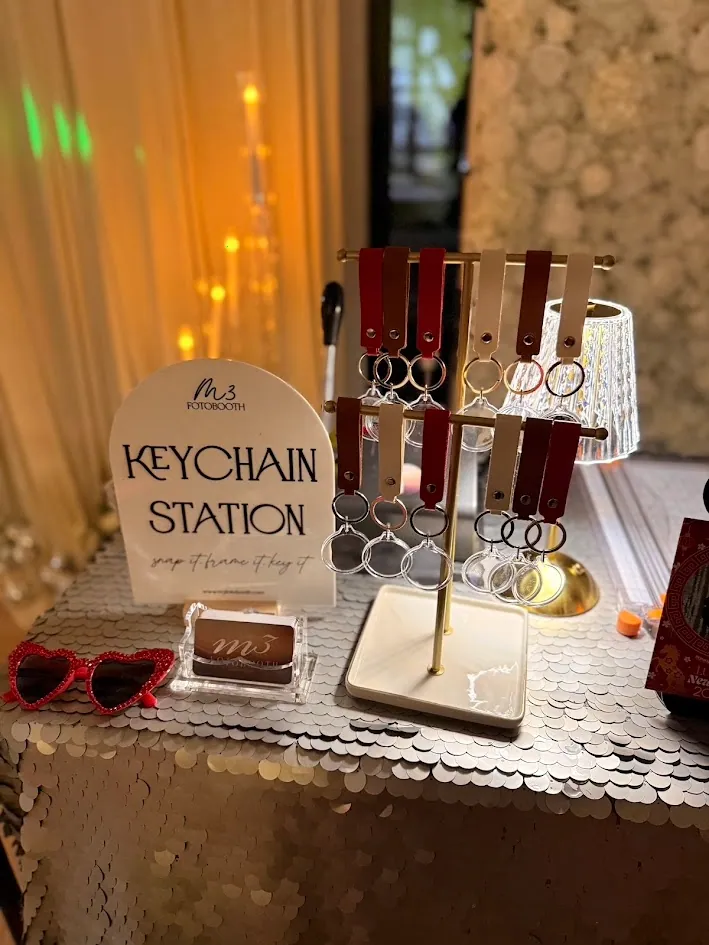 A keychain station sign on a sequin cloth beside a gold rack of finished leather keychains and a pair of red heart shaped sunglasses