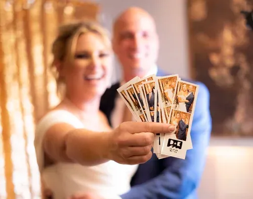 A couple holding a fan of just printed photo strips out toward the camera, warm light behind them