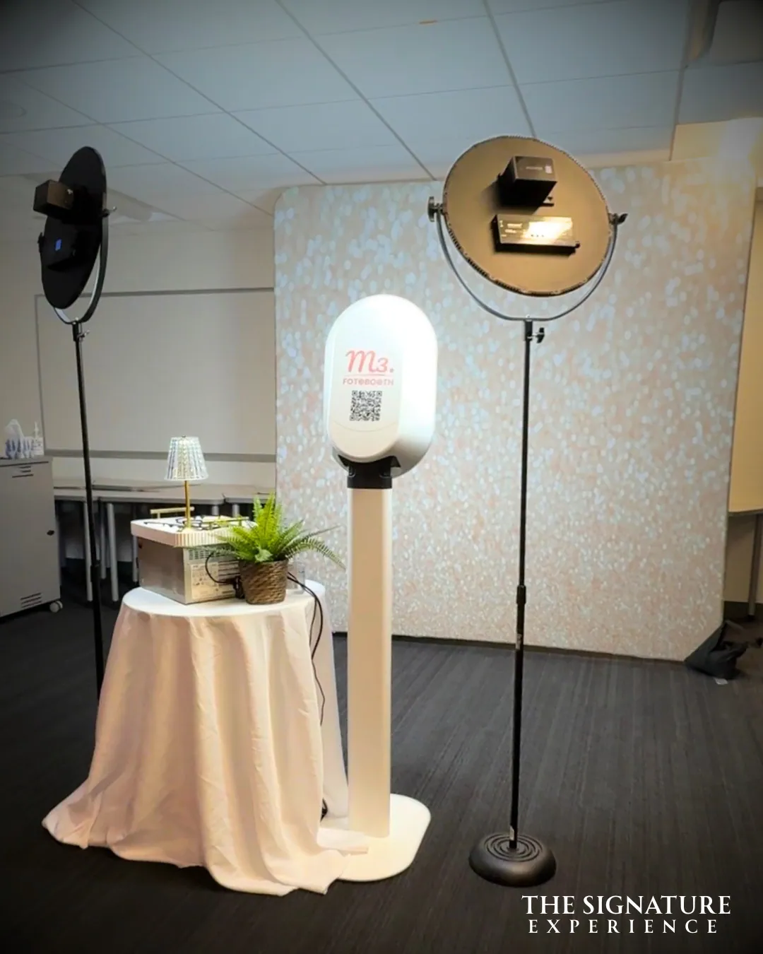 A white photo booth on its stand beside a cream draped table set with a small lamp and a fern, a ring light on a stand alongside