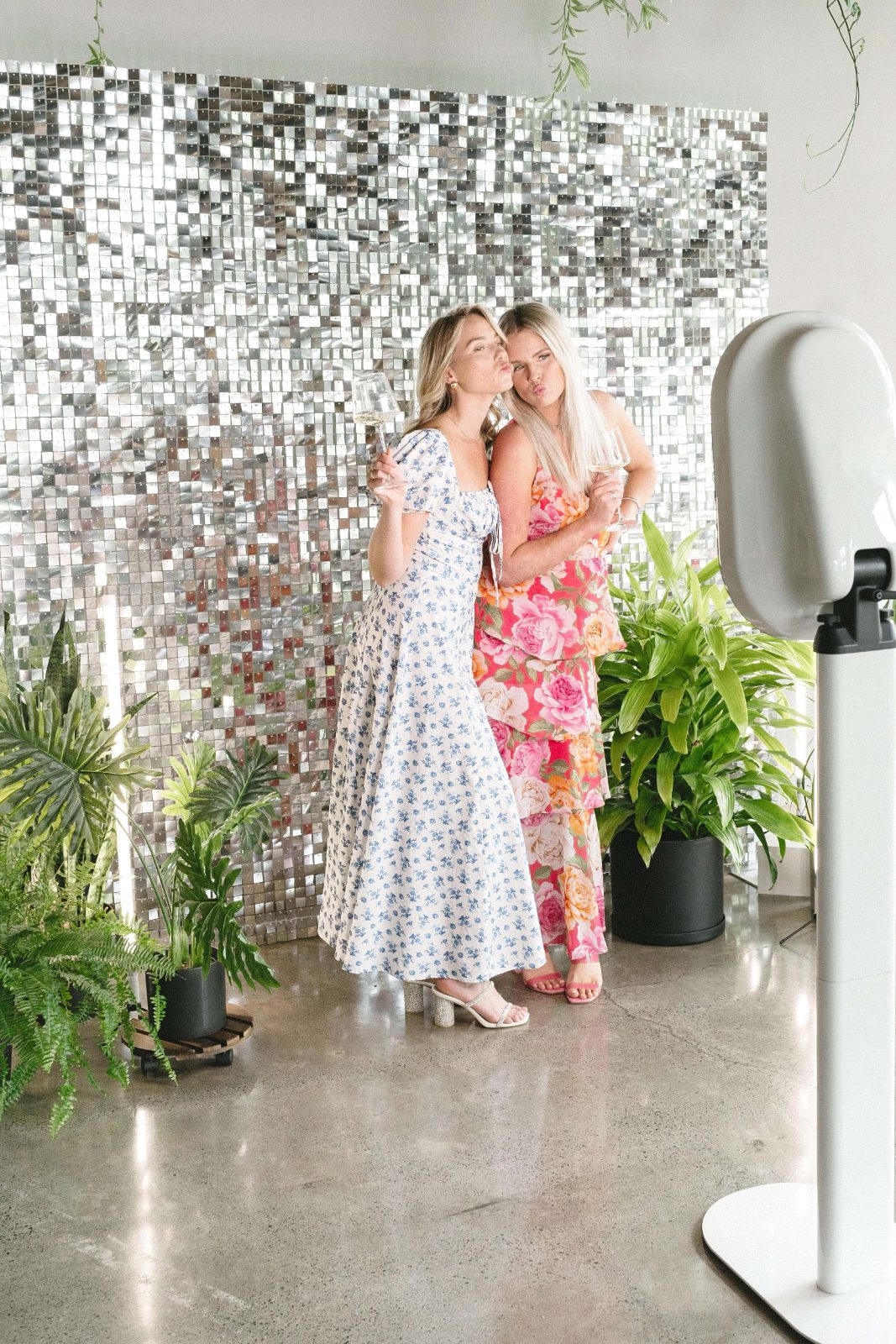 Two guests in floral dresses pose before a silver sequin wall next to a white photo booth