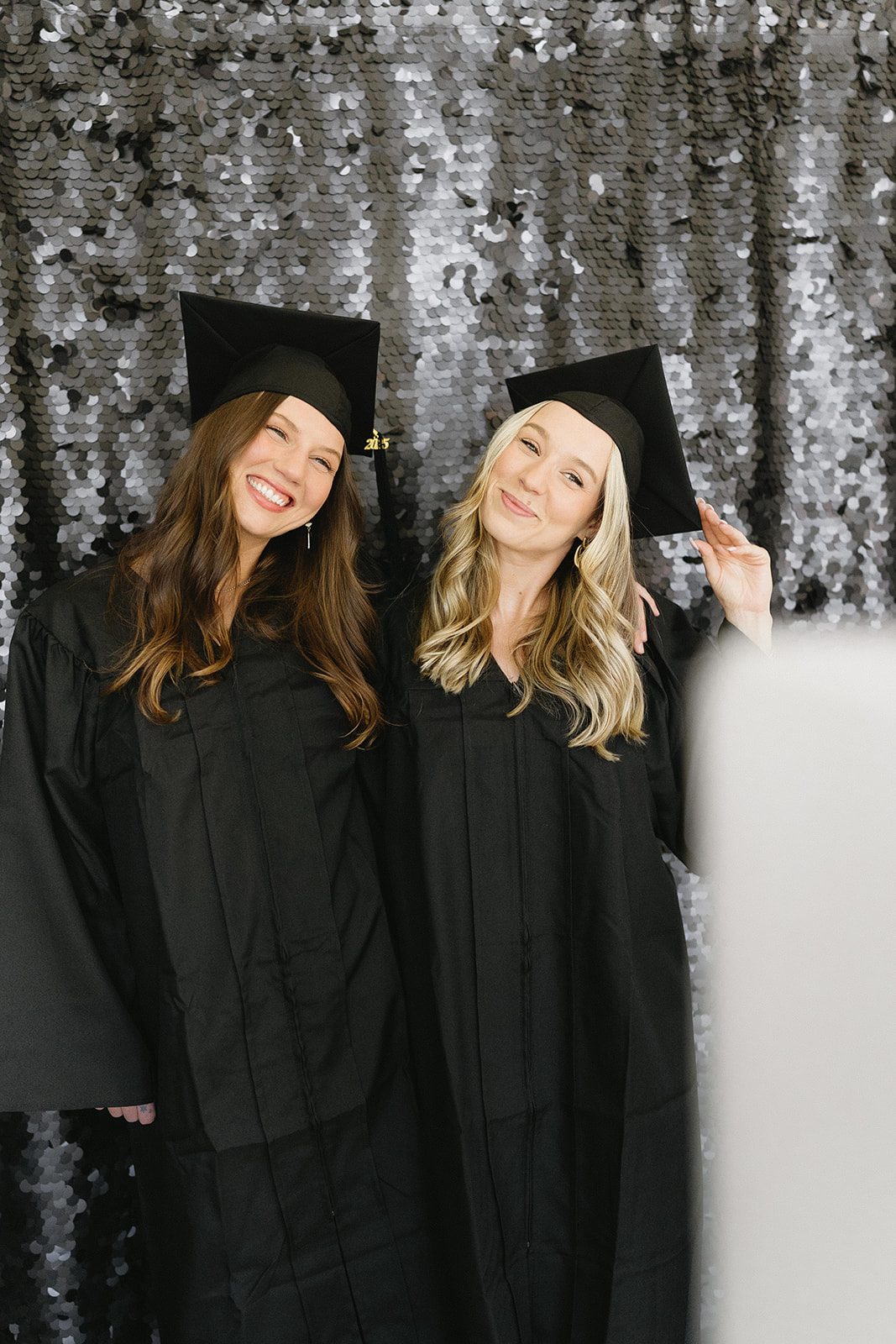 Two graduates in caps and gowns posing together in front of a silver sequin backdrop