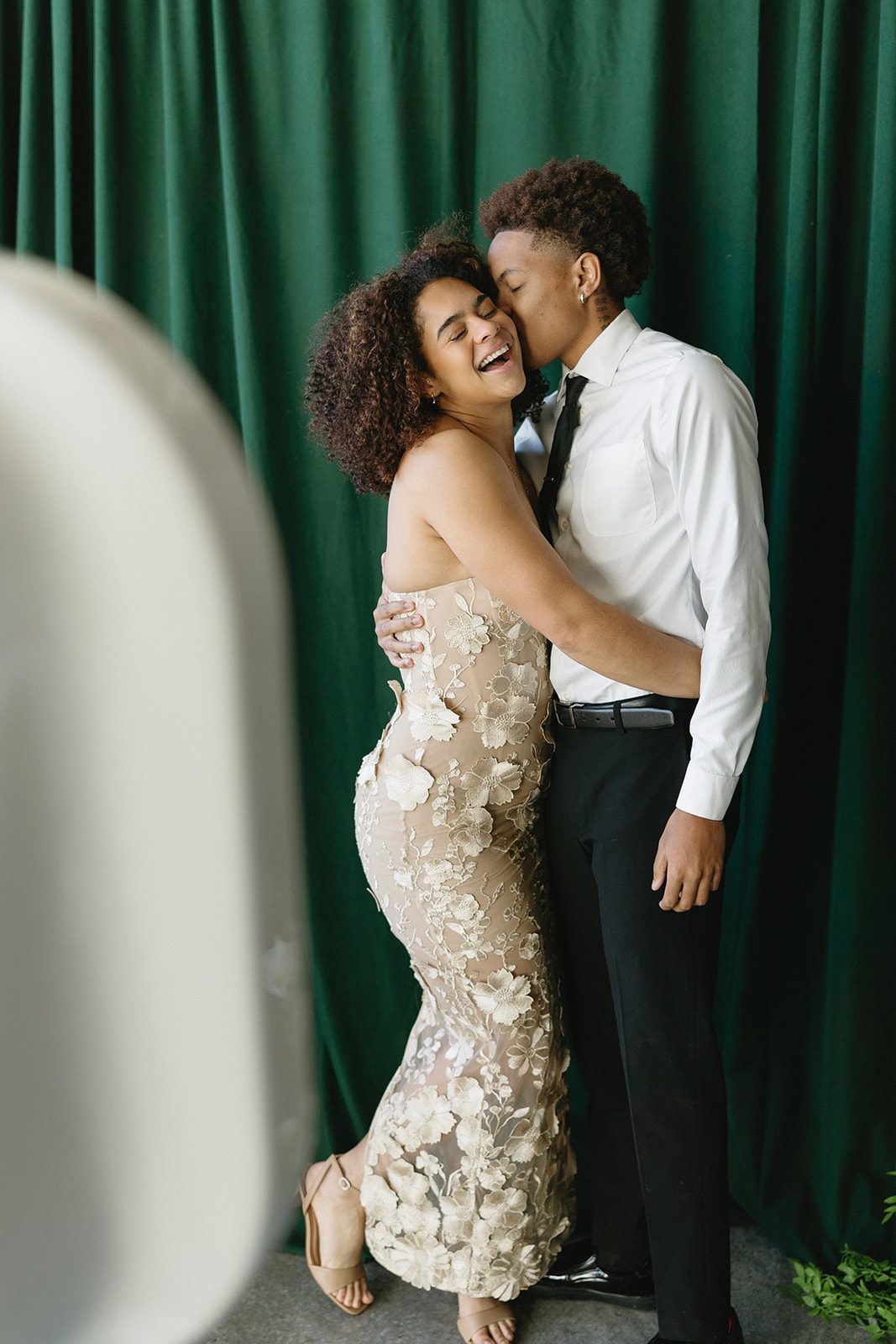 A couple laughing mid kiss in front of a deep green curtain, the edge of the booth just in frame