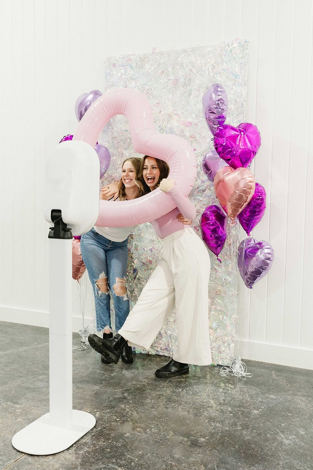 Two guests laugh inside a giant inflatable heart prop at a white photo booth with heart balloons