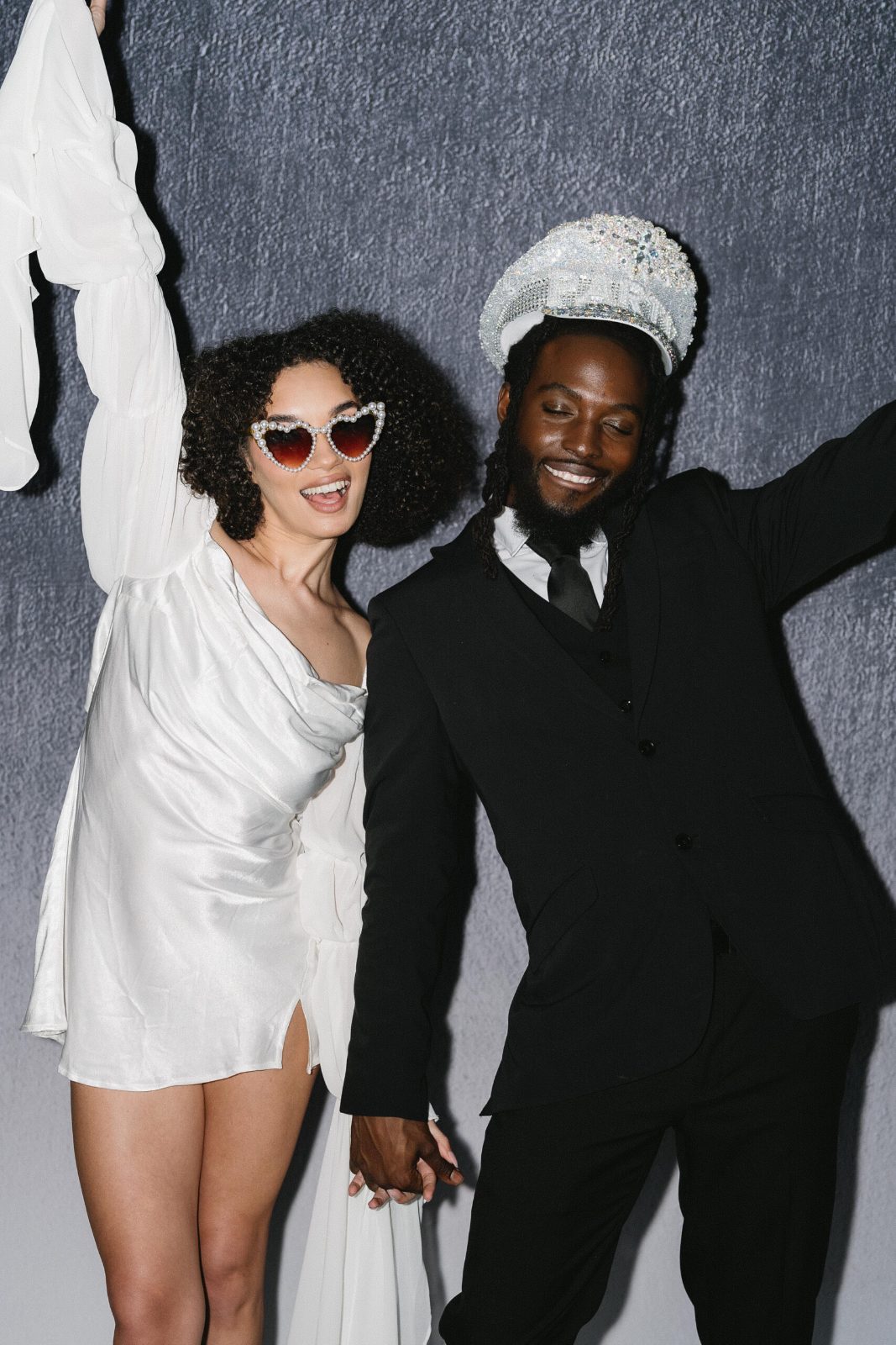 A couple in a white dress and black suit dance, she in heart sunglasses, he in a sparkly captain's hat
