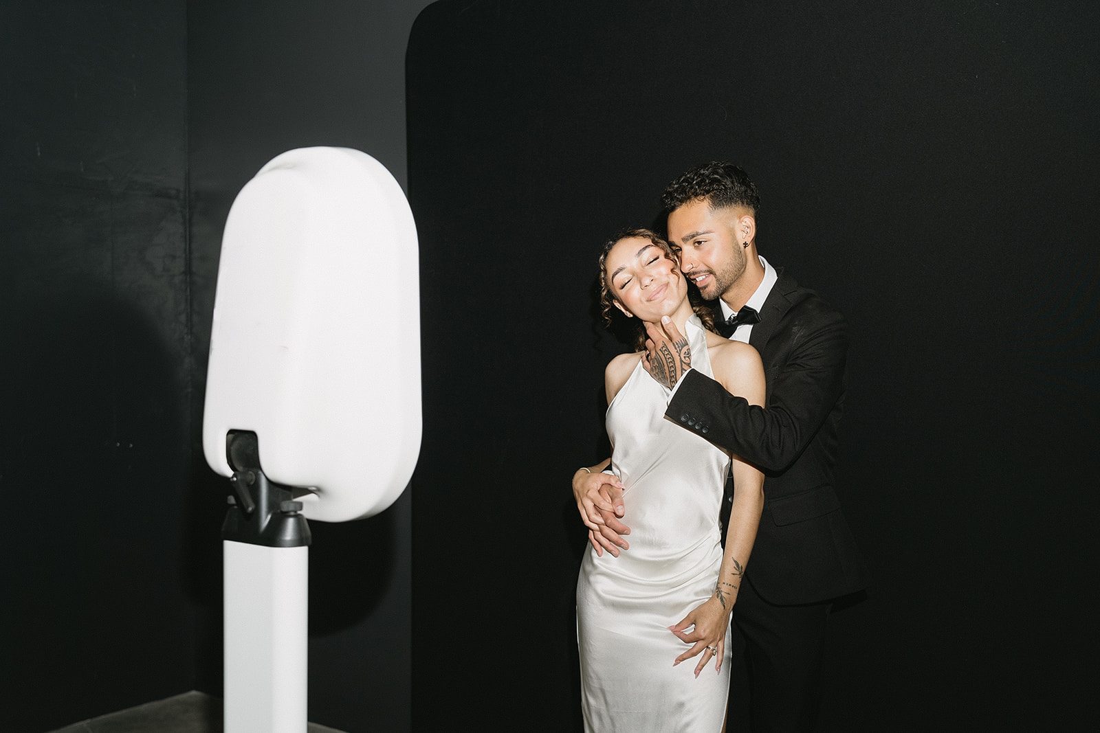 A couple in a white satin gown and a black tuxedo leaning into each other against a black backdrop, lit from the left by the booth