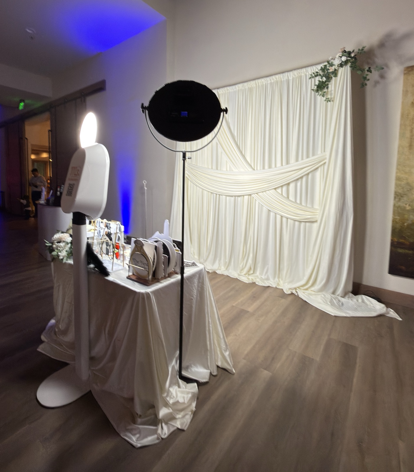 The booth beside a draped prop table in front of white curtains and greenery, lit blue from above