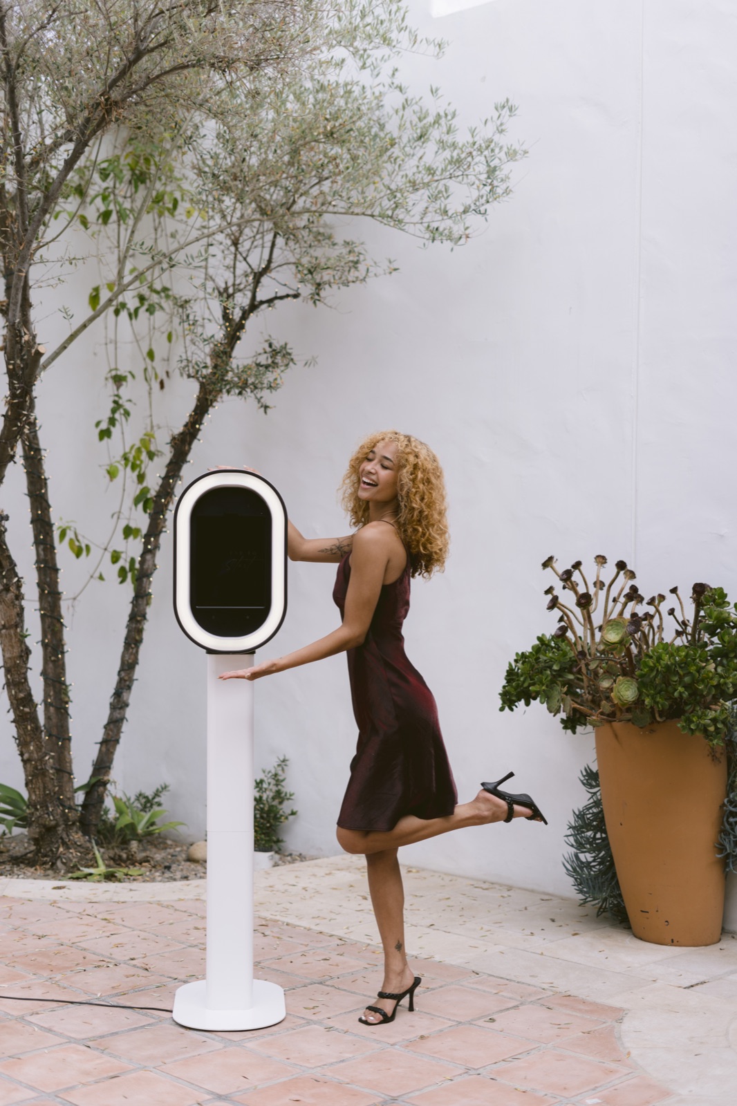 A guest in a burgundy satin dress leaning on the booth outdoors, beside a potted tree against a white wall