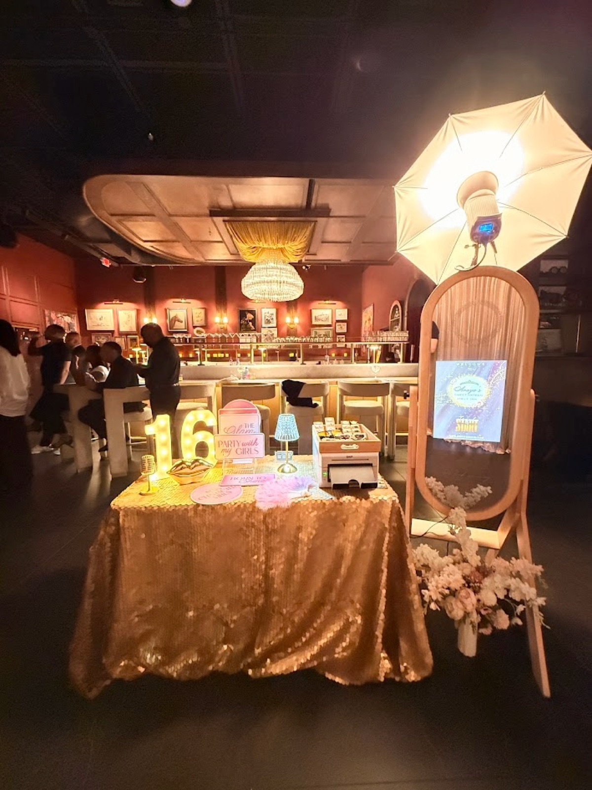 Mirror photo booth and umbrella light beside a gold sequin table with a lit 16 marquee and printer