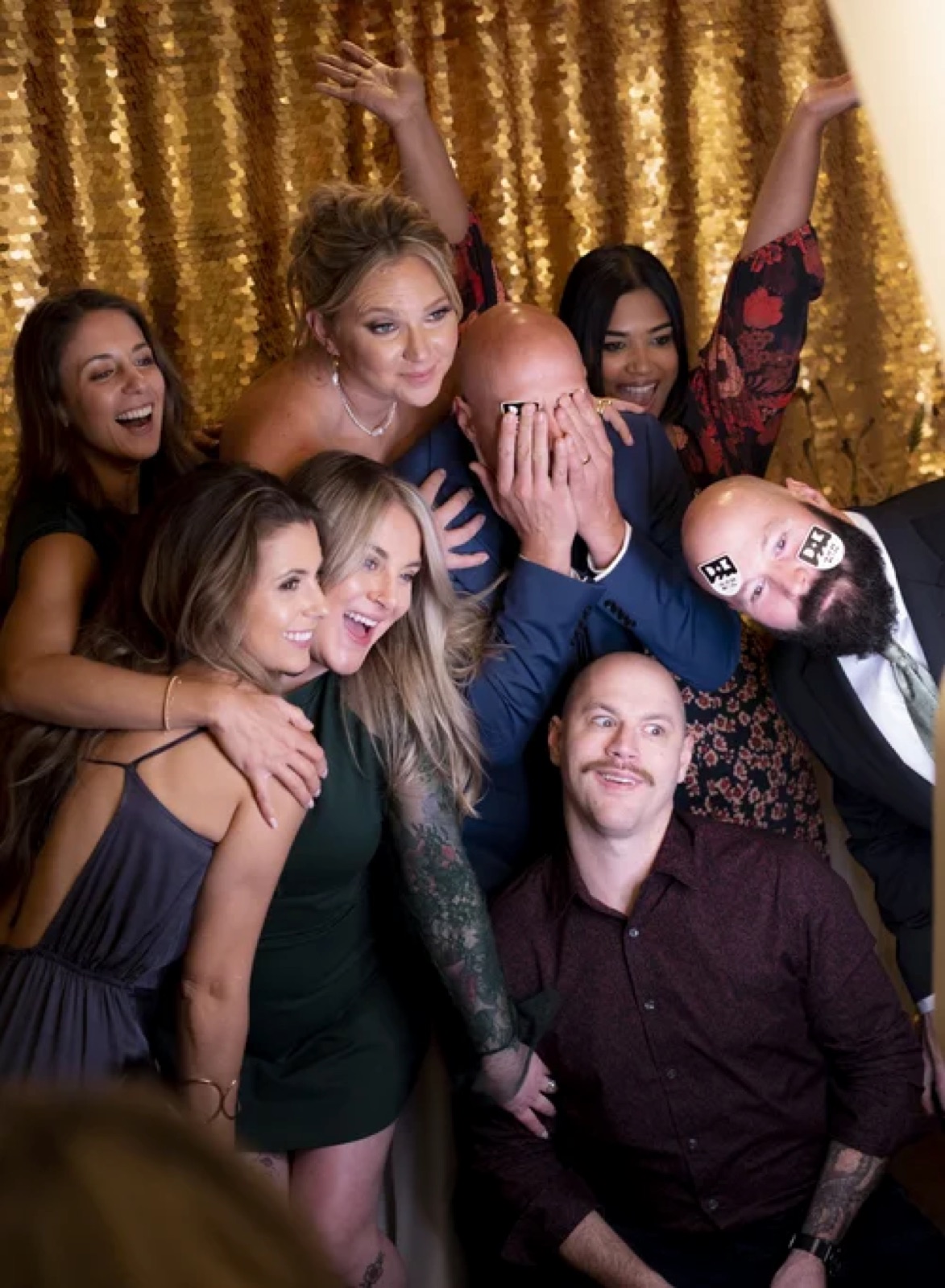 A crowd of guests piled into the frame together in front of a gold sequin backdrop, arms in the air