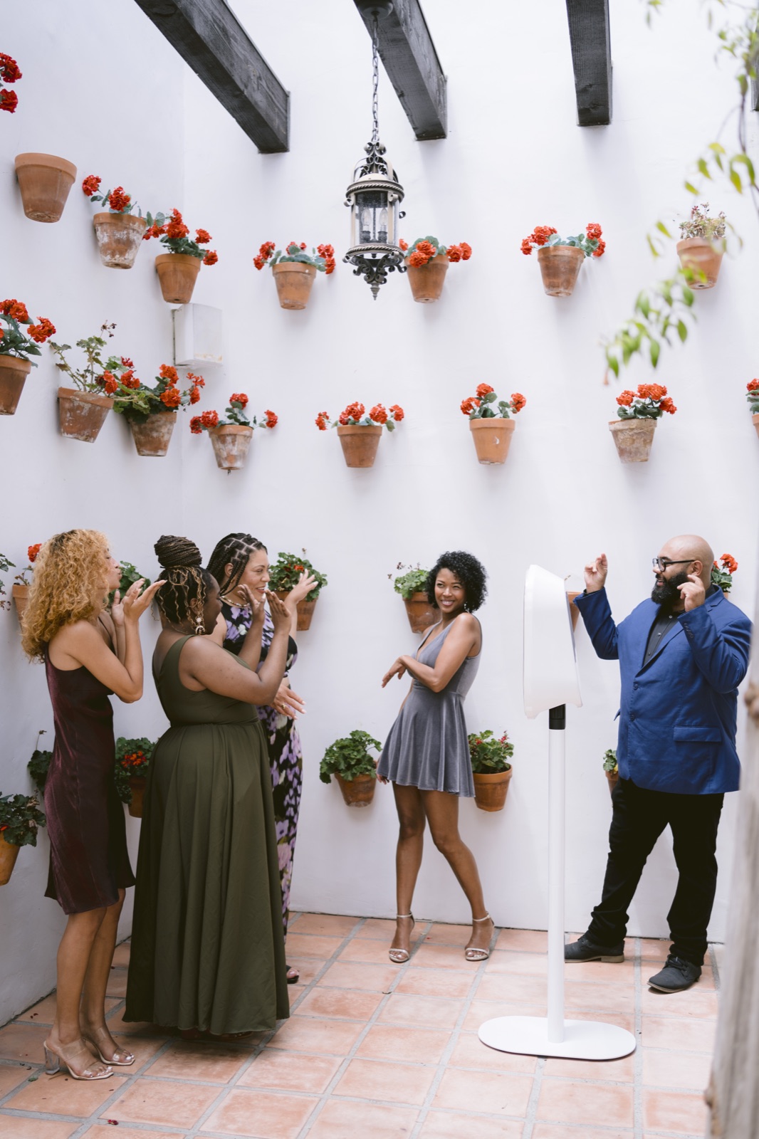 Three guests clap as a woman in a gray dress poses at a white photo booth among potted geraniums
