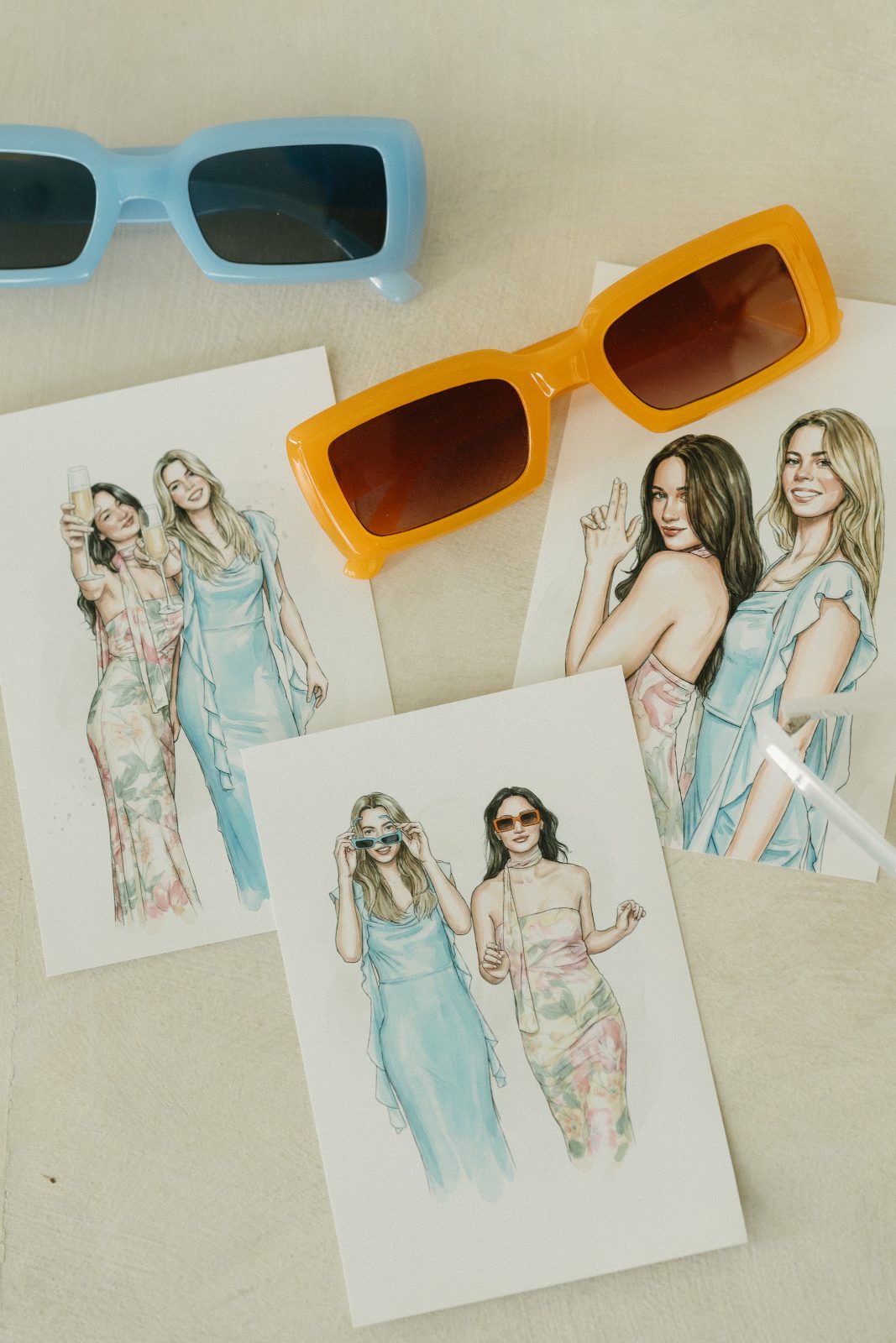 Three watercolor portrait prints of guests laid out on a table with two pairs of colored sunglasses