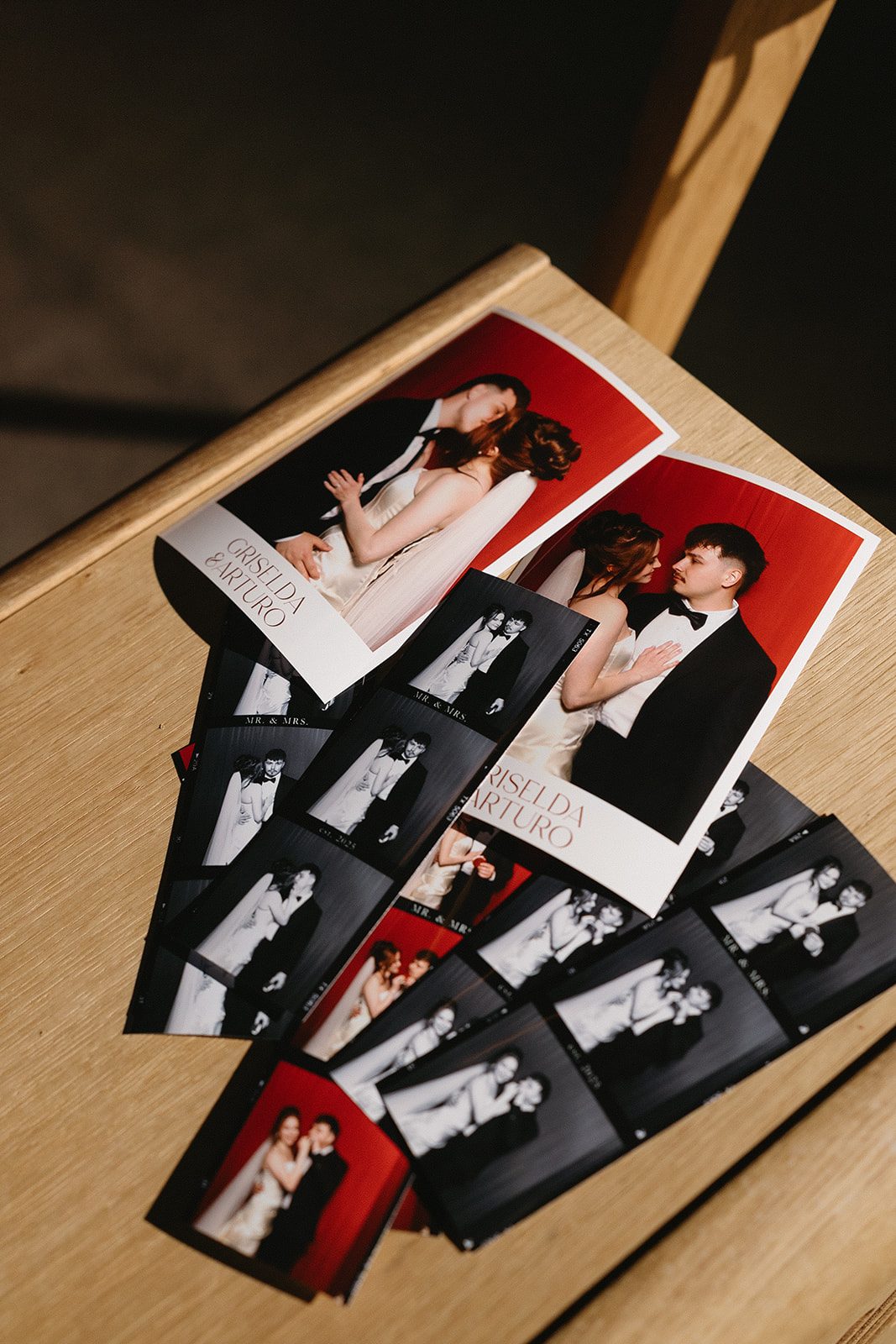 Finished photo strips scattered across a wood table, some printed on deep red and some in black and white, each carrying the couple's names