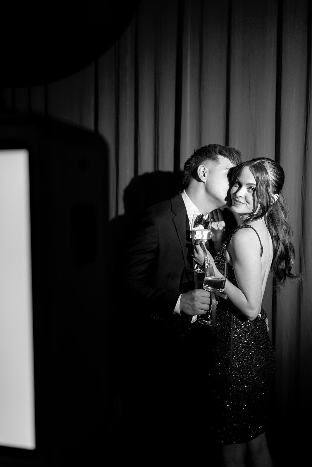 A black and white frame of a couple holding champagne glasses, him kissing her cheek, against a dark curtain