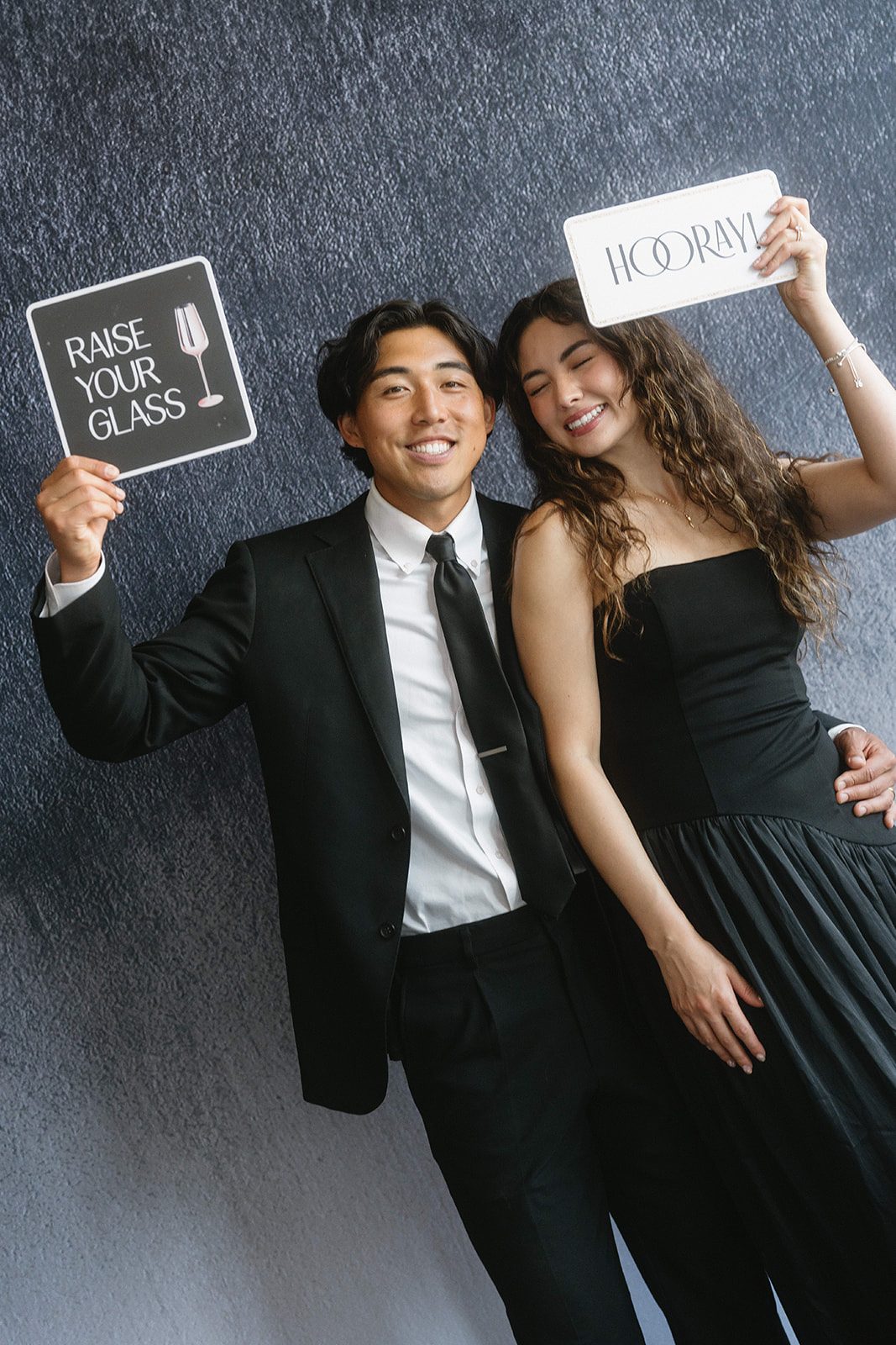 Two guests in black tie holding up Raise Your Glass and Hooray signs against a dark textured backdrop
