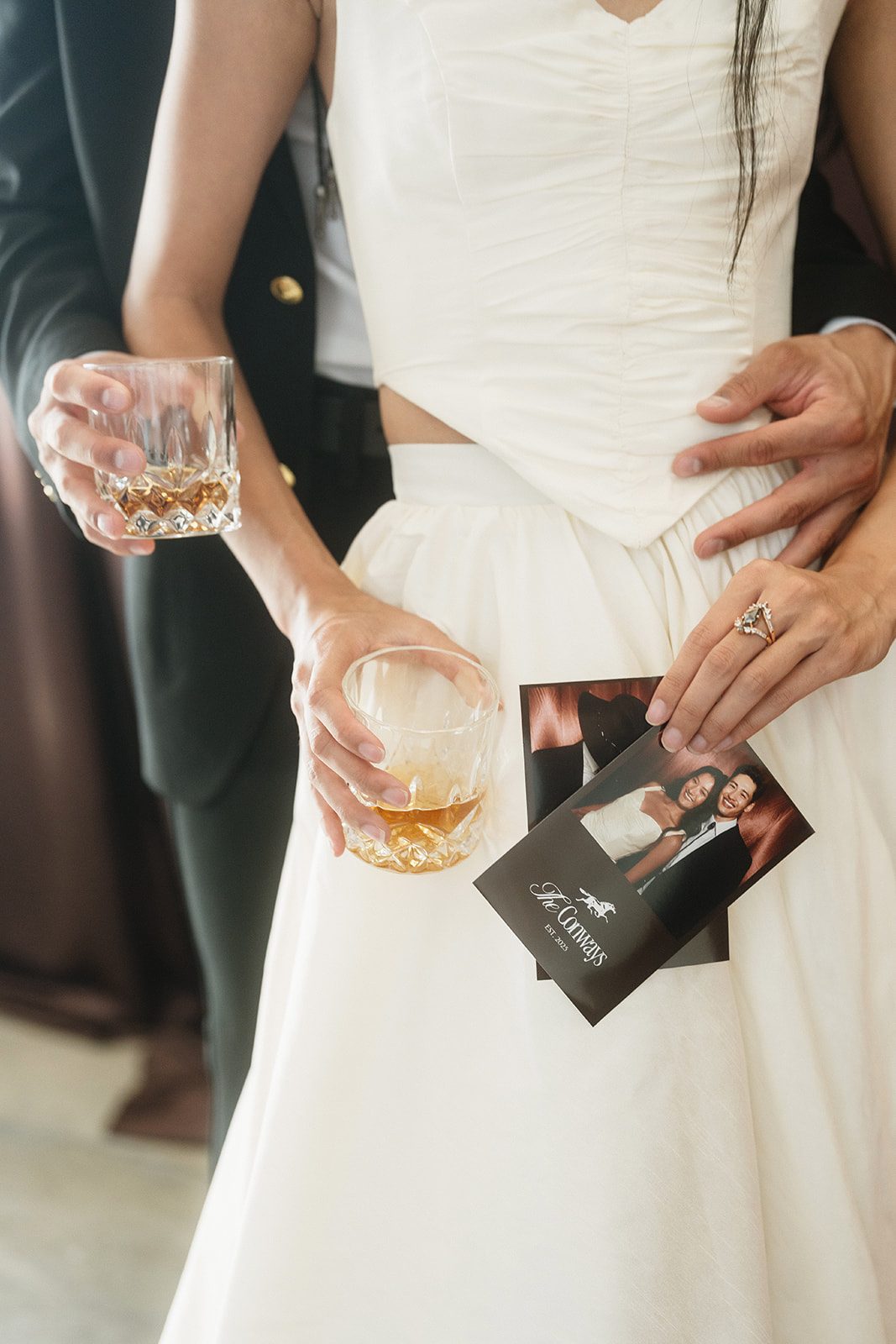 A woman in white holding a whiskey glass and photo booth prints, a man's hand at her waist